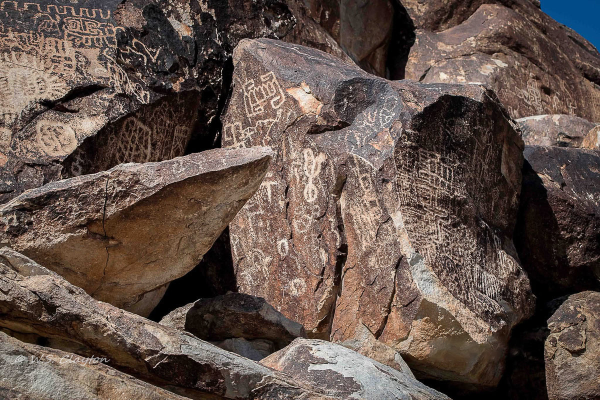 Rock formations with engraved petroglyph carvings and symbols, photographed outdoors in bright sunlight against a small patch of blue sky.