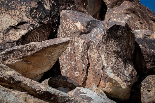 Rock formations with engraved petroglyph carvings and symbols, photographed outdoors in bright sunlight against a small patch of blue sky.