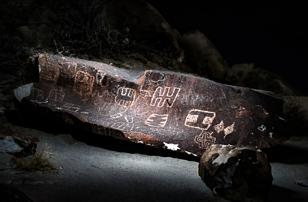 A dramatic low-light photograph of a large rock slab with ancient petroglyph carvings, illuminated by a flashlight against a dark, rocky background.